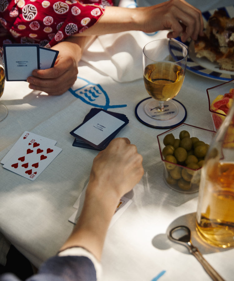 Three people enjoy a lively evening playing cards at a table with wine, olives, snacks, and food on a white-and-blue cloth, using PORTA Playing Cards.