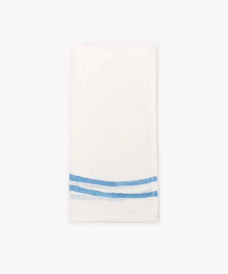 The Onda Napkin, Set of 6, features folded white linen napkins hand block-printed with two parallel blue wave-like stripes near one edge, displayed on a plain light background.