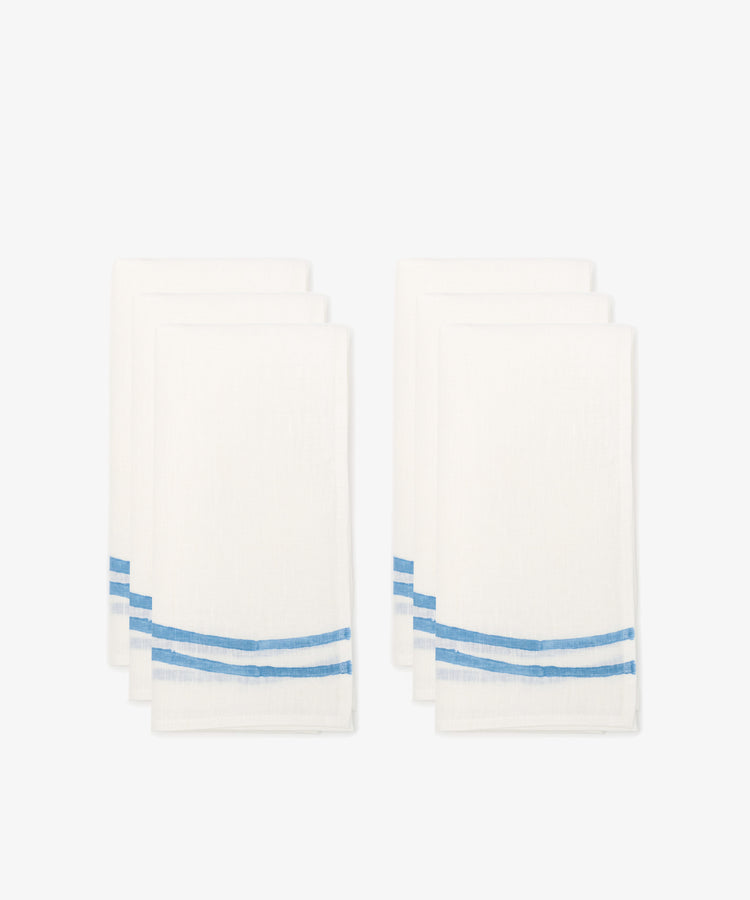 Six Onda napkins, white linen with two blue wave-like stripes near one edge, arranged in two stacks against a plain white background.