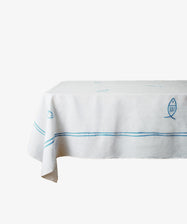 The Pezza Tablecloth features white hand block-printed fabric with blue fish motifs and stripes, shown draped over a rectangular table against a plain white background.