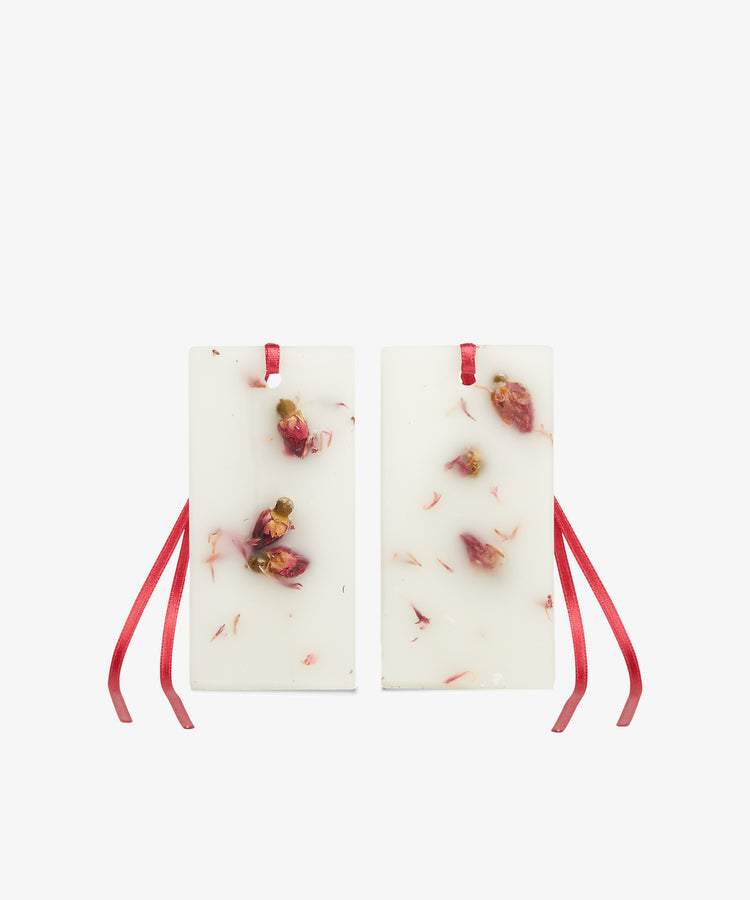 The Rosa Novella Scented Wax Tablets, Set of 2, are rectangular white wax tablets with embedded dried rose petals and red ribbon loops at the top.