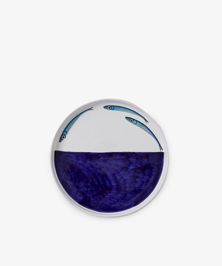 The Tommaso Flying Fish Platter is a round ceramic serving piece with a deep blue section inspired by Amalfi Coast waters and two illustrated flying fish above the blue area on a white background.