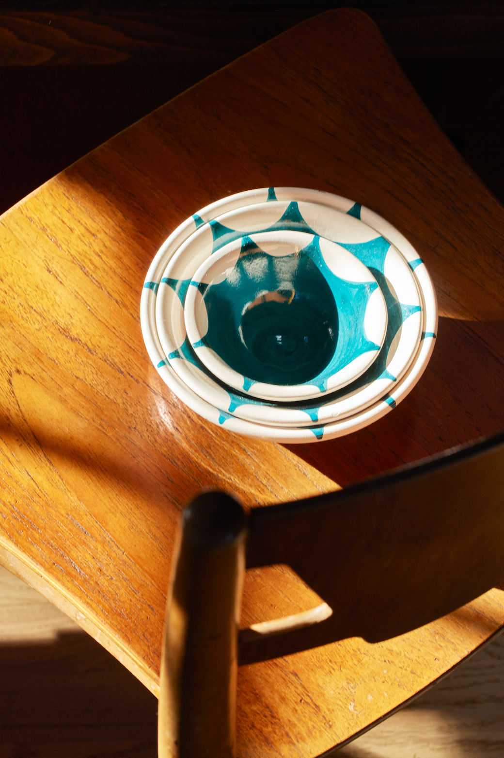 The Stella Nesting Bowls, featuring a handmade turquoise and white geometric design, rest on a wooden chair in sunlight—ideal for anyone who loves unique kitchenware.