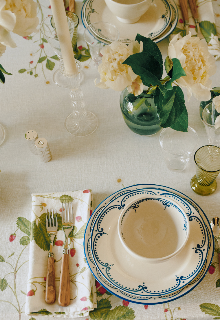 A table set with patterned plates, bowls, cutlery on a napkin, hand-blown glassware, the Elsa Candleholder, and vases with white flowers atop a floral tablecloth.