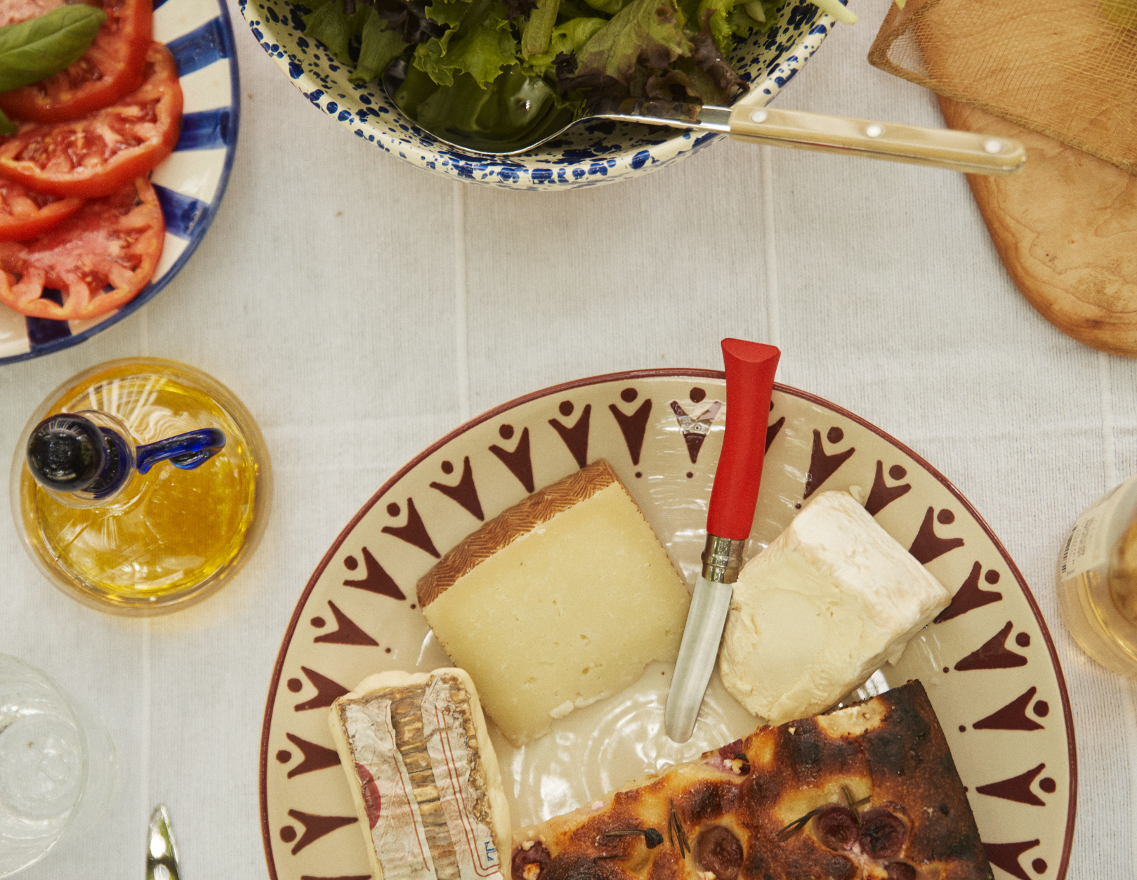 A table set with assorted cheeses, sliced tomatoes, leafy greens, olive oil, bread, and the Primo Kids Knife on a white tablecloth.