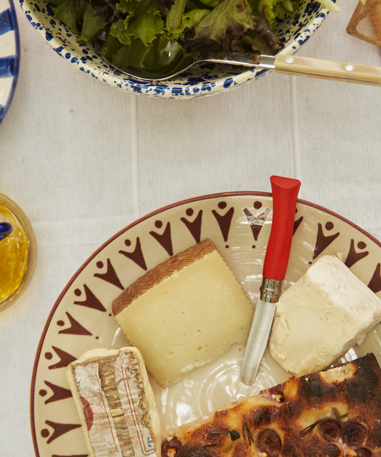 A table set with assorted cheeses, sliced tomatoes, leafy greens, olive oil, bread, and the Primo Kids Knife on a white tablecloth.