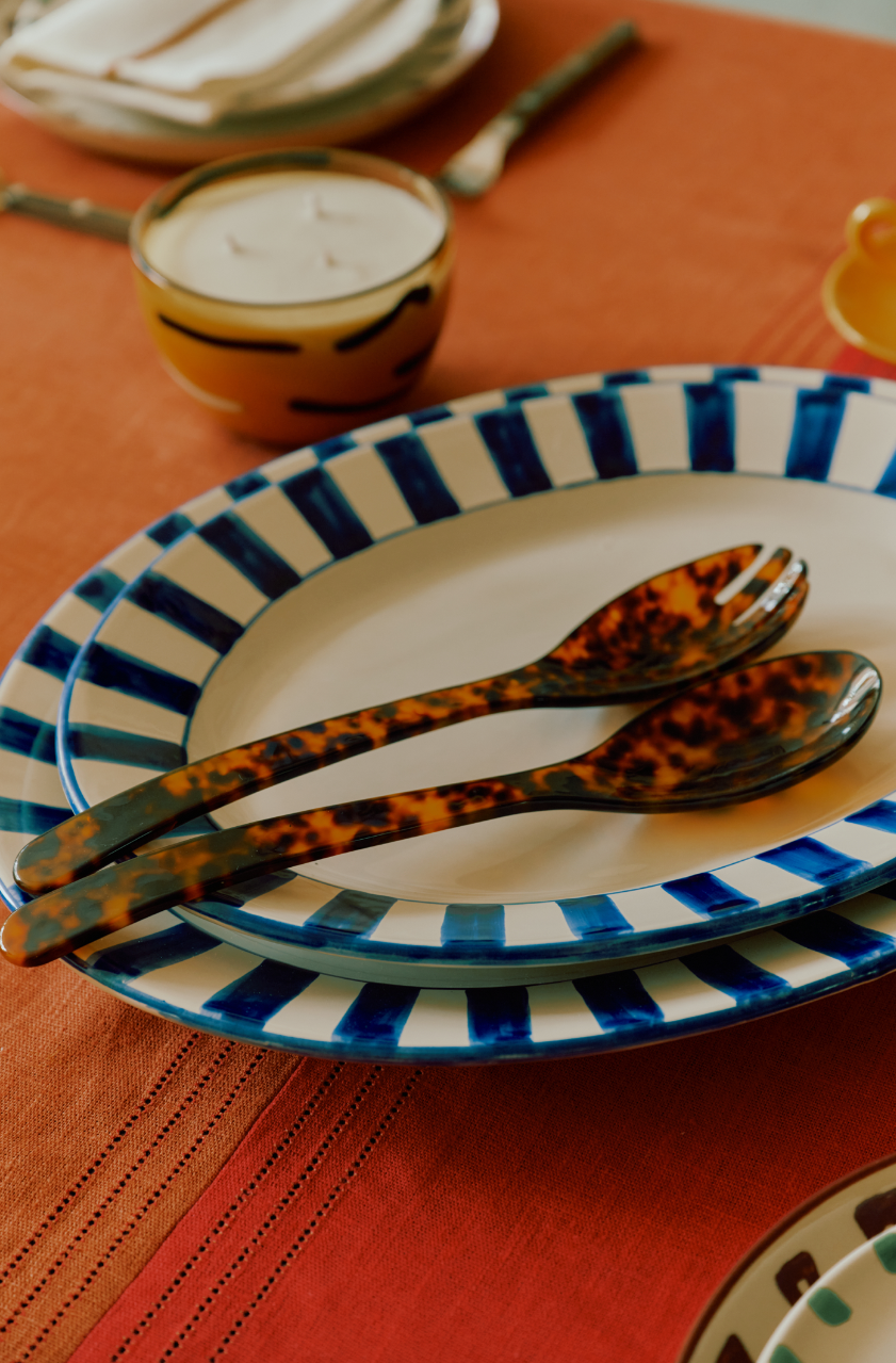 The Bestia Serving Set sits on an orange tablecloth beside two blue and white striped plates, a yellow-rimmed bowl, and a candle.