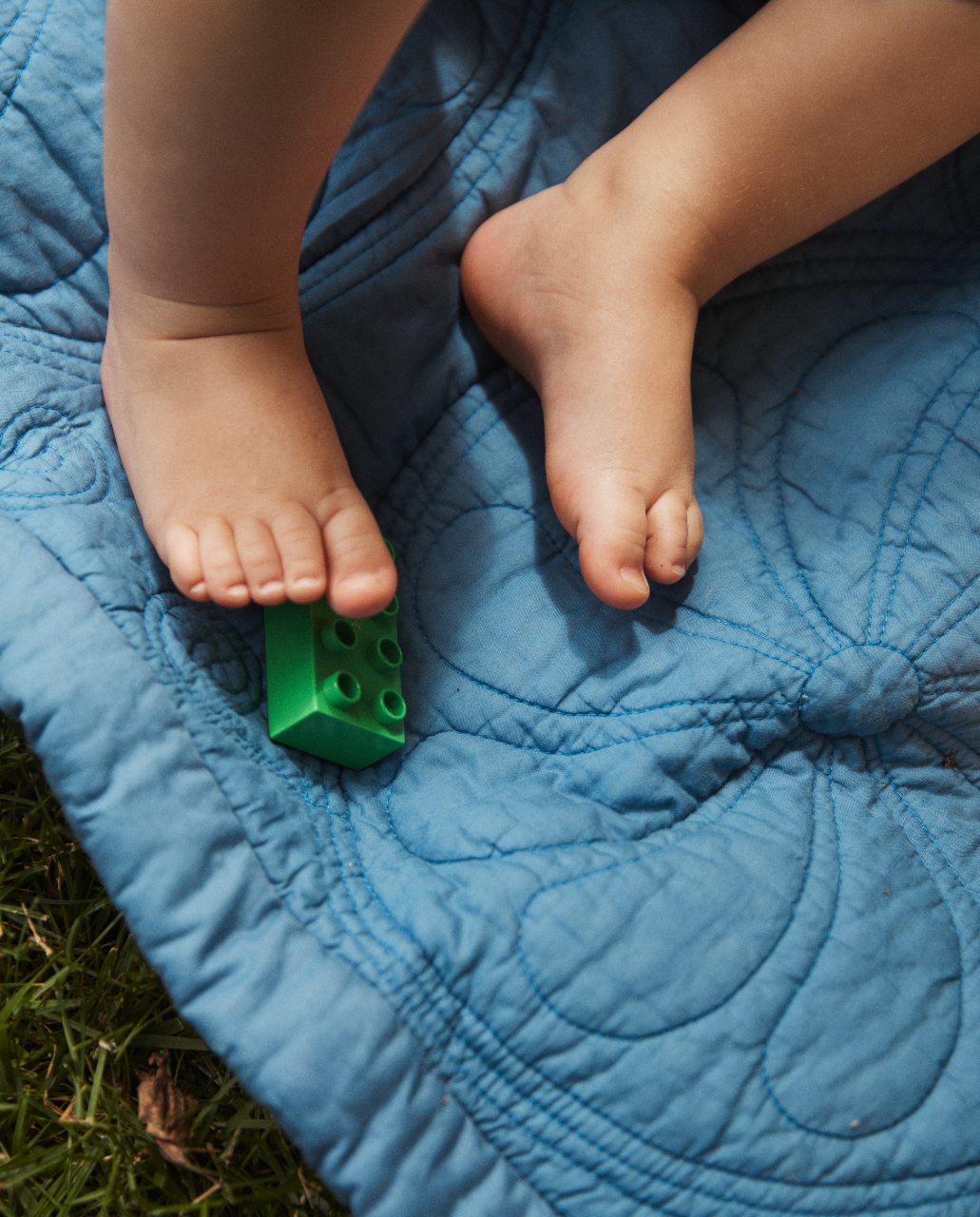 A childs bare feet rest on the Dawn Twin Bedspread, a blue double-sided quilt, with a green toy block under one foot, on grass.