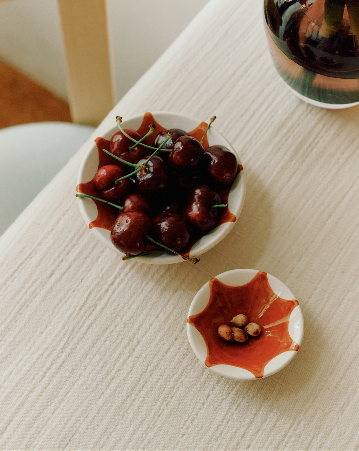 A bowl of fresh cherries in one of our Stella Nesting Bowls sits next to a small dish for cherry pits on a light textured tablecloth—perfect for aperitivo snacks or experimenting with creative presentation.