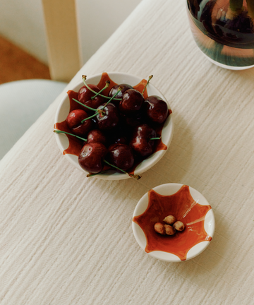 A bowl of fresh cherries in one of our Stella Nesting Bowls sits next to a small dish for cherry pits on a light textured tablecloth—perfect for aperitivo snacks or experimenting with creative presentation.