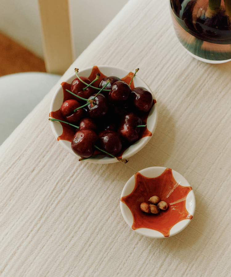 A bowl of fresh cherries in one of our Stella Nesting Bowls sits next to a small dish for cherry pits on a light textured tablecloth—perfect for aperitivo snacks or experimenting with creative presentation.