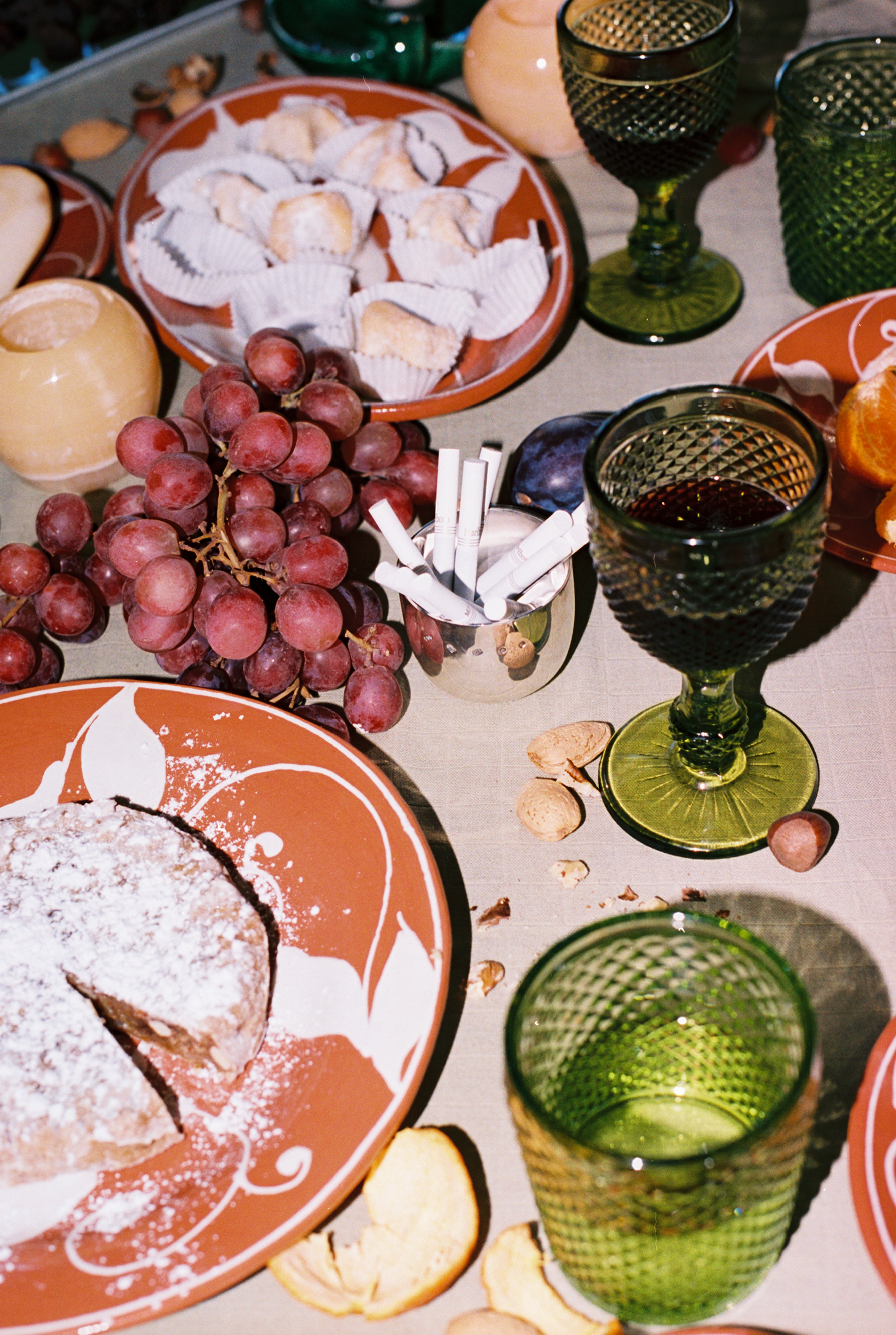 A table set with Celeste Red Wine Glasses, red grapes, assorted nuts, sweets, a powdered cake, and a cup holding cigarettes highlights subtle coloring and sophistication.