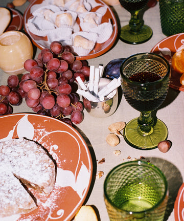 A table set with Celeste Red Wine Glasses, red grapes, assorted nuts, sweets, a powdered cake, and a cup holding cigarettes highlights subtle coloring and sophistication.