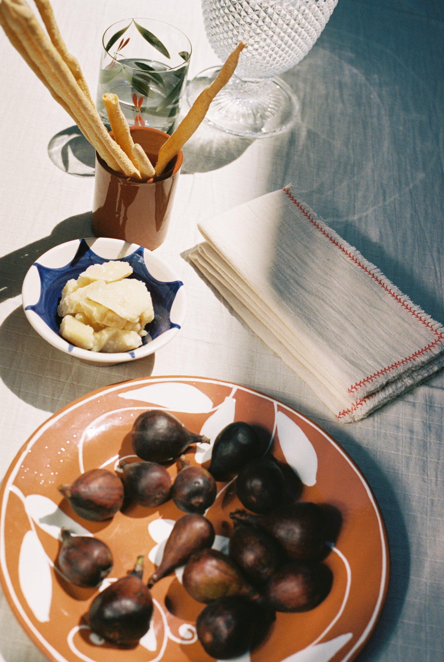 A tabletop scene features breadsticks in a cup, water glasses, neatly stacked Filial Napkin, Set of 6 cotton napkins, a bowl of cheese, and fresh figs on a patterned plate.