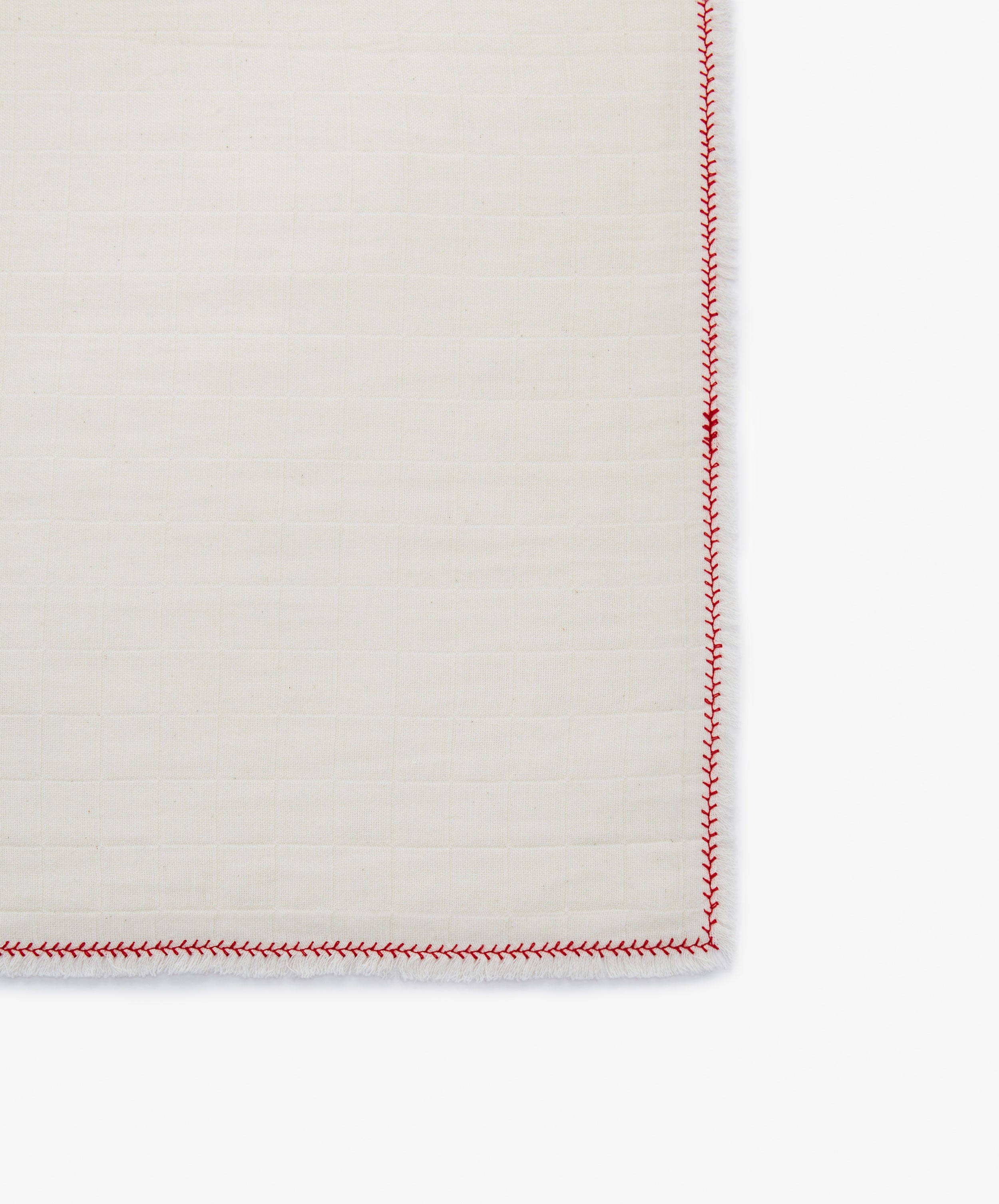 A close-up of the corner of a Filial Napkin, Set of 6, in cotton cream with a grid pattern and red zigzag edge stitching, displayed on a tabletop.