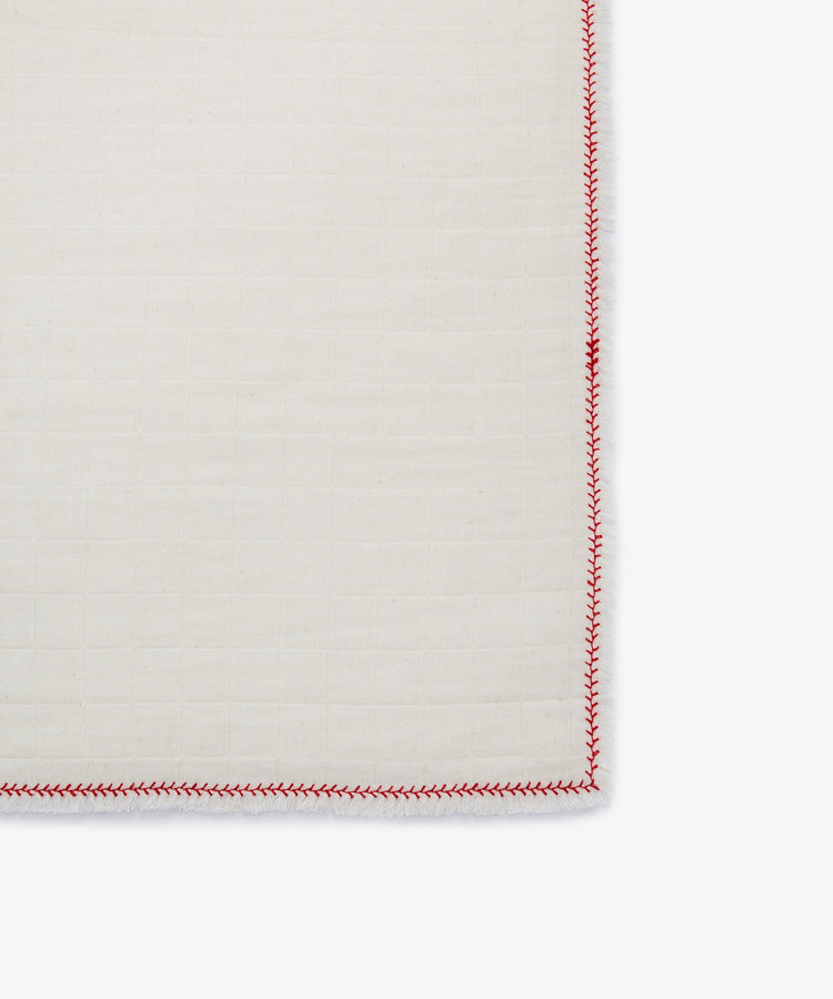 A close-up of the corner of a Filial Napkin, Set of 6, in cotton cream with a grid pattern and red zigzag edge stitching, displayed on a tabletop.