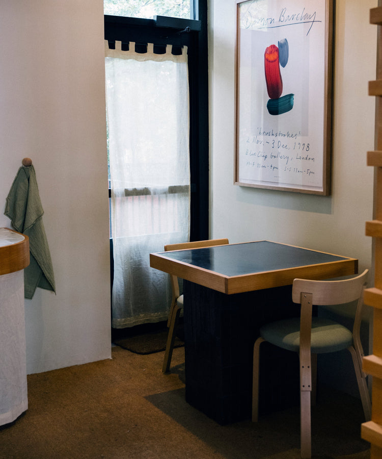 Small dining table with two chairs beside a wall, featuring a large framed art print and a window with sheer curtains.