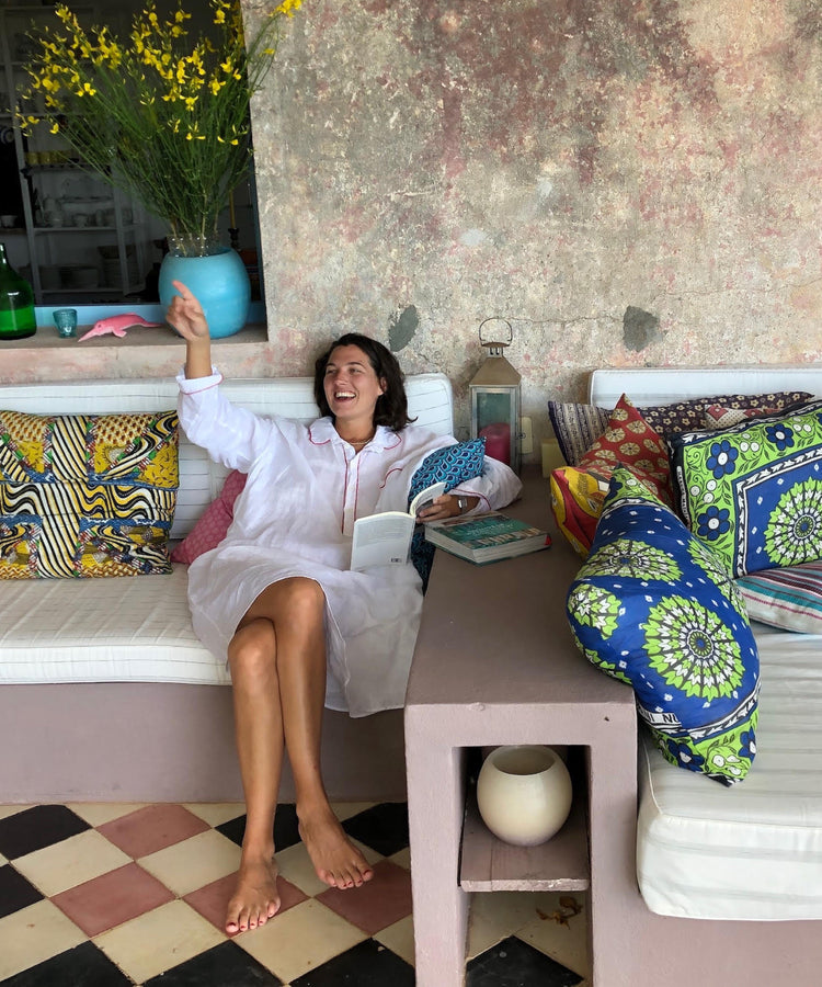 Woman sitting on a cushioned bench with colorful pillows, smiling and holding a book.