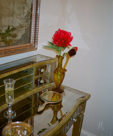 A mirrored table holds an Alba Vase—a yellow, hand-blown glass piece—with a red flower, a glass of drink, a round handheld mirror, and a small gold dish under a framed painting.