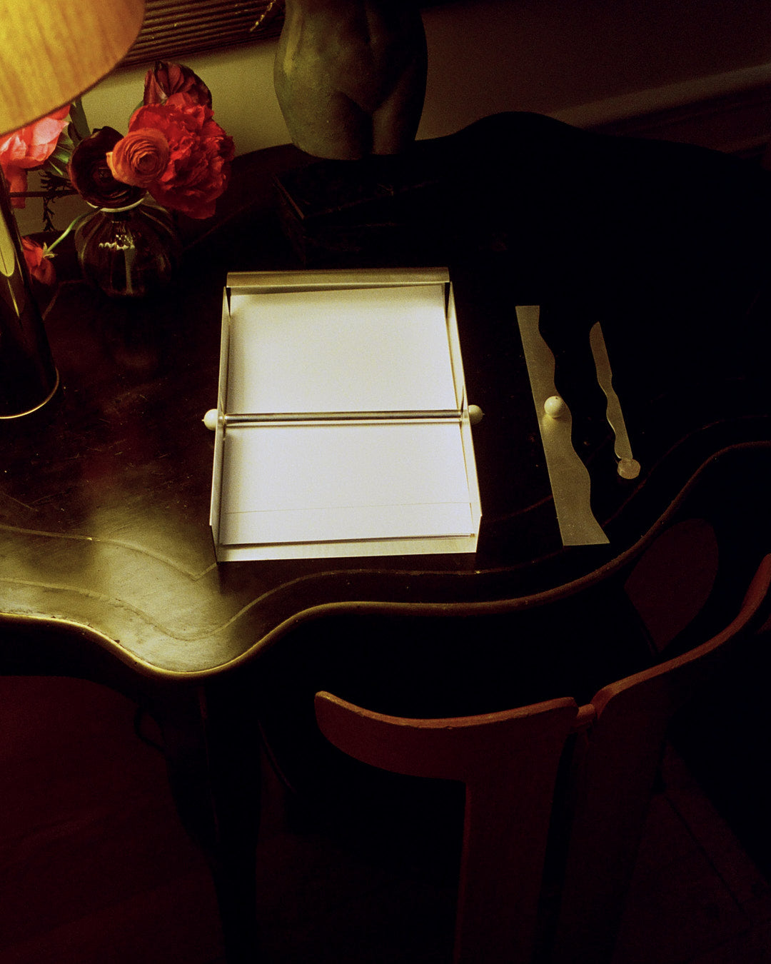 A dark wooden desk with a white open box, a lamp, a vase of red flowers, and decorative objects on top—including the Paper Holder, a silver-plated brass rectangular accessory that adds a modern touch.