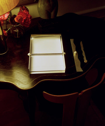 A dark wooden desk with a white open box, a lamp, a vase of red flowers, and decorative objects on top—including the Paper Holder, a silver-plated brass rectangular accessory that adds a modern touch.