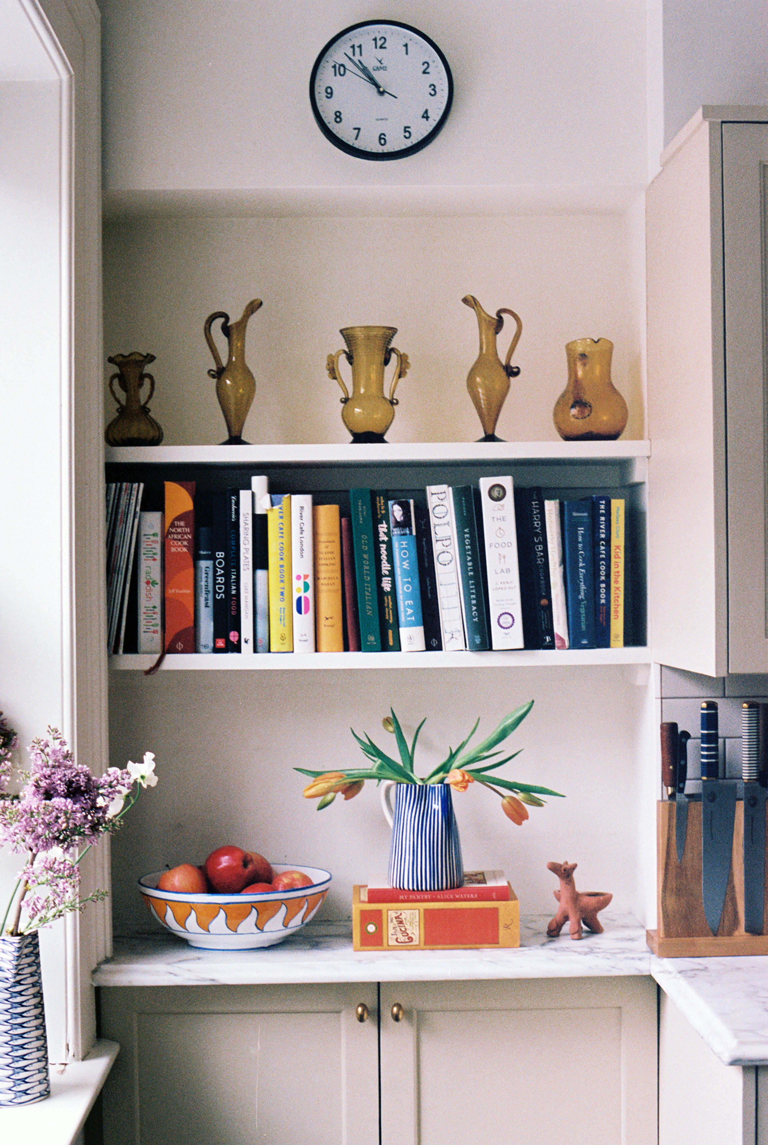 A kitchen shelf with yellow vases, a clock, books, the Palio Serving Bowl, The Unicorn filled with oranges, a vase of tulips, a knife block, and a small deer figurine on a marble countertop evokes the charm of Siena, Italy.