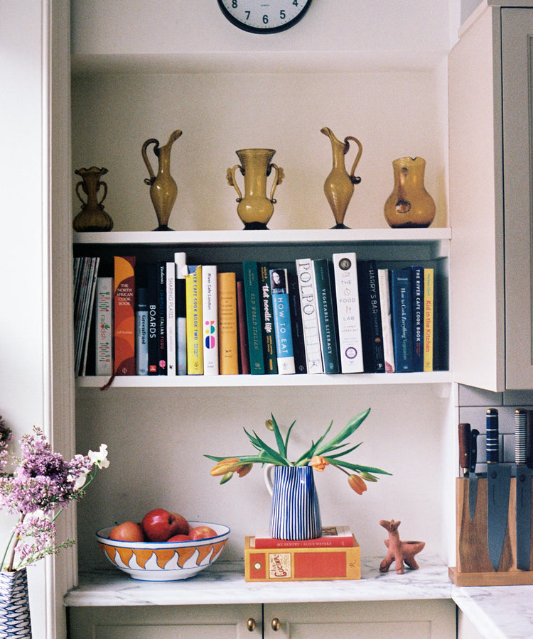 A kitchen shelf with yellow vases, a clock, books, the Palio Serving Bowl, The Unicorn filled with oranges, a vase of tulips, a knife block, and a small deer figurine on a marble countertop evokes the charm of Siena, Italy.