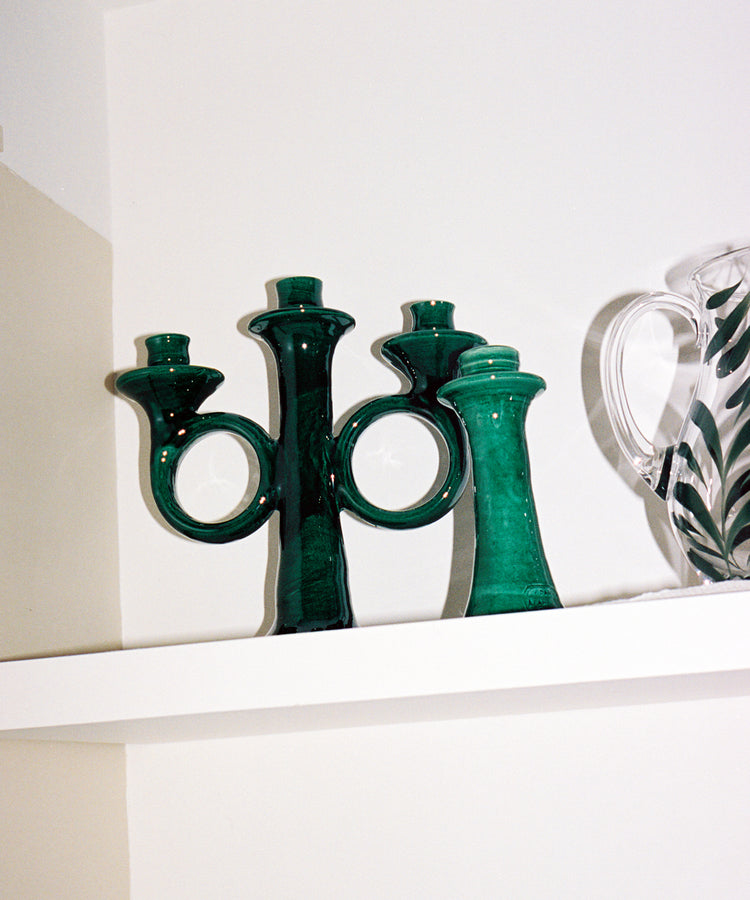 The Loretta Candelabra and a handmade glass pitcher with green leaf patterns sit on a white shelf against a white wall.
