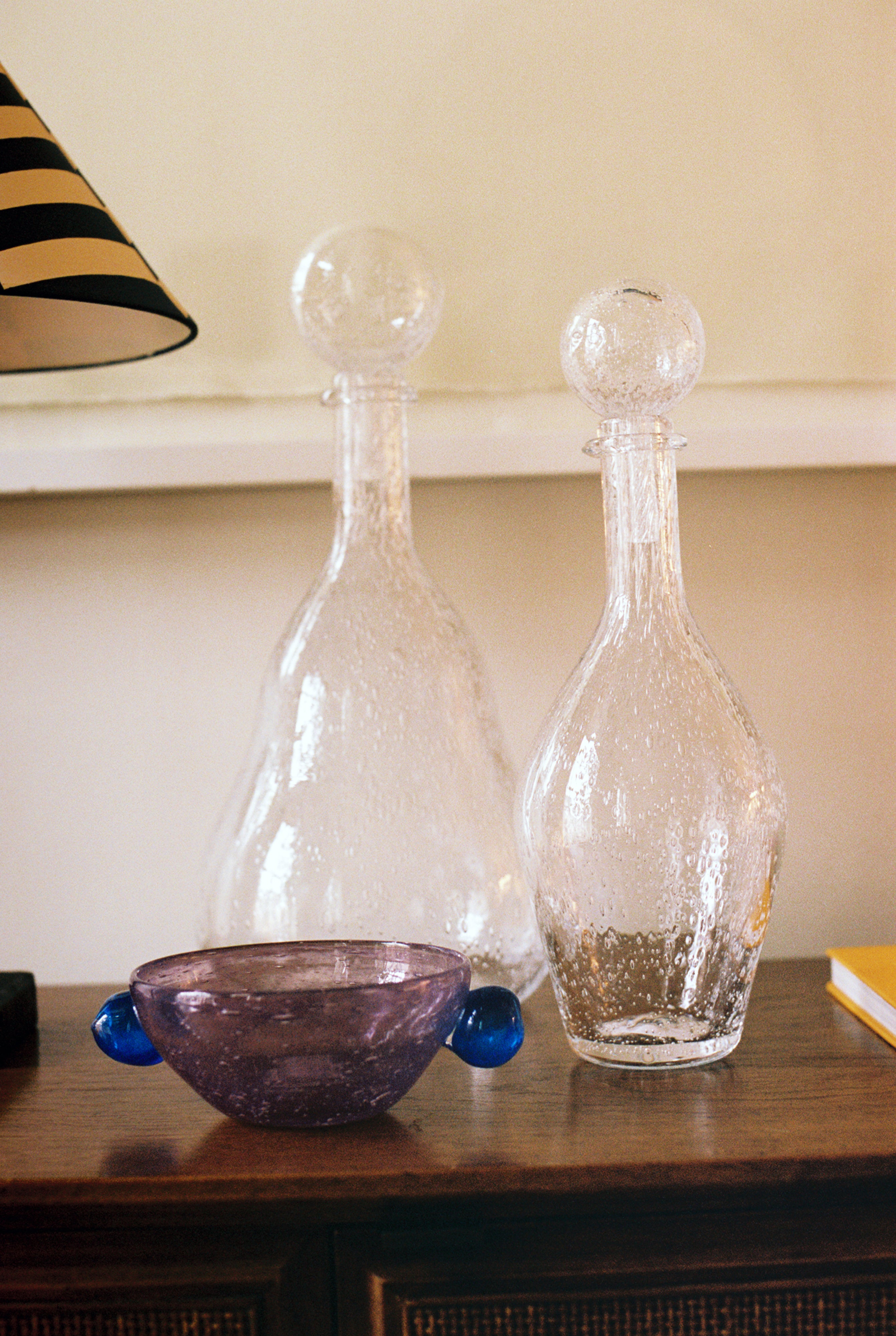 The Pavot Snack Bowl, featuring a purple handmade design with blue handles, is displayed on a wooden surface beside two clear glass decanters with round stoppers, near a lamp and a yellow book.