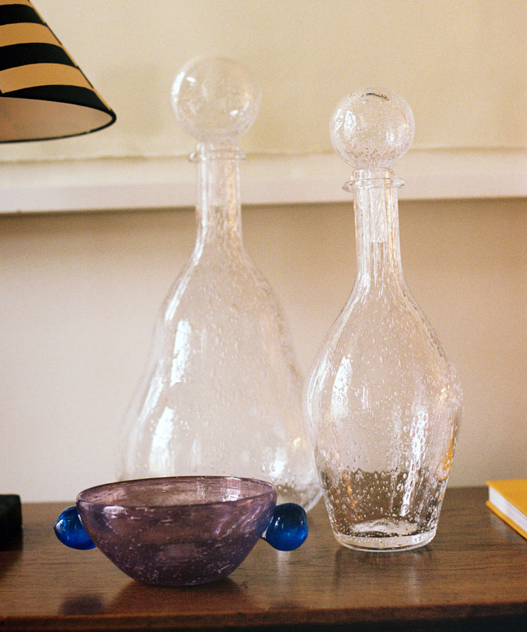 The Pavot Snack Bowl, featuring a purple handmade design with blue handles, is displayed on a wooden surface beside two clear glass decanters with round stoppers, near a lamp and a yellow book.