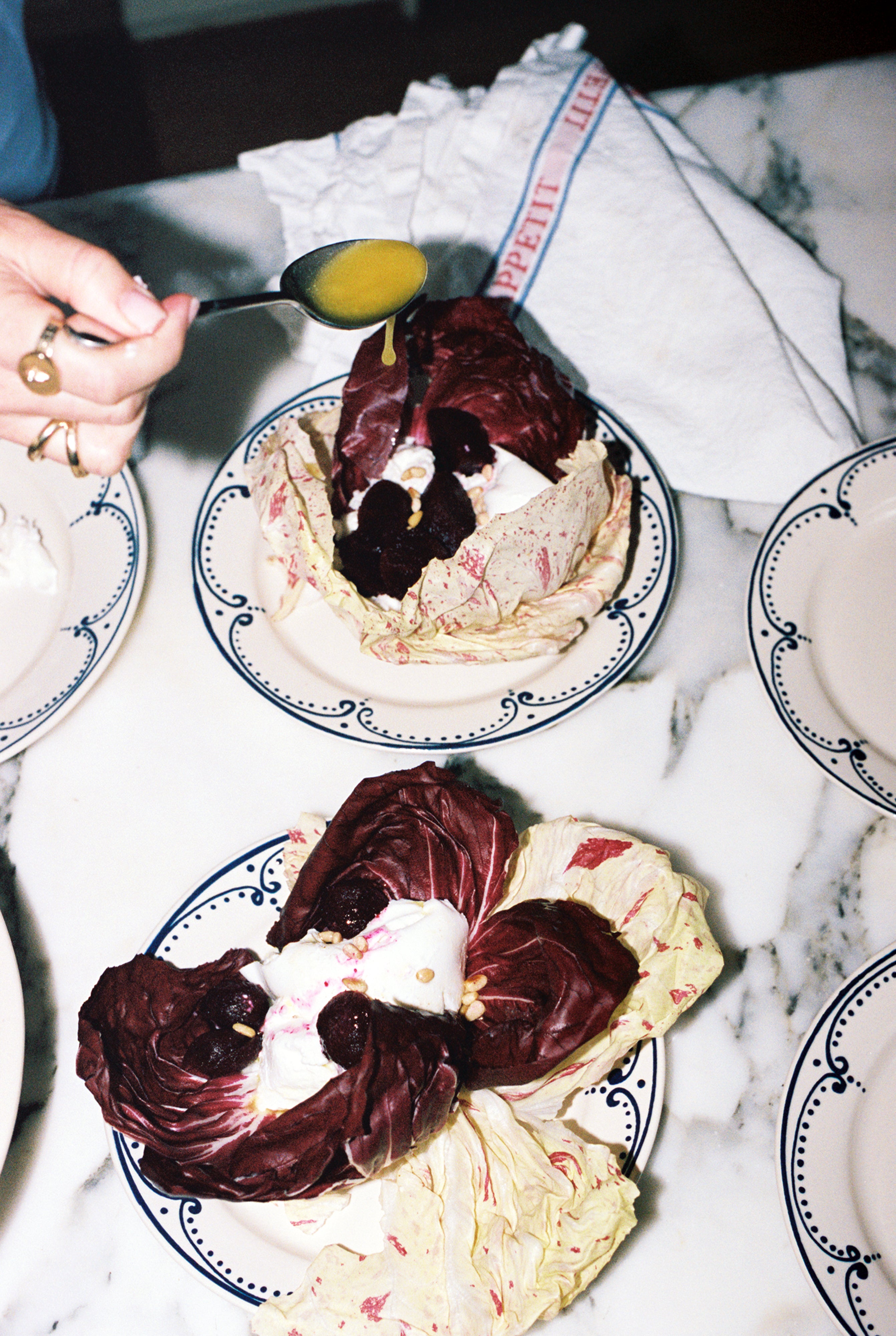 A hand drizzles yellow dressing over radicchio leaves filled with beets and cream, served on the Arco Small Plate, Set of 4, featuring a traditional design, placed on a marble surface with a napkin and other plates.