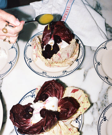 A hand drizzles yellow dressing over radicchio leaves filled with beets and cream, served on the Arco Small Plate, Set of 4, featuring a traditional design, placed on a marble surface with a napkin and other plates.