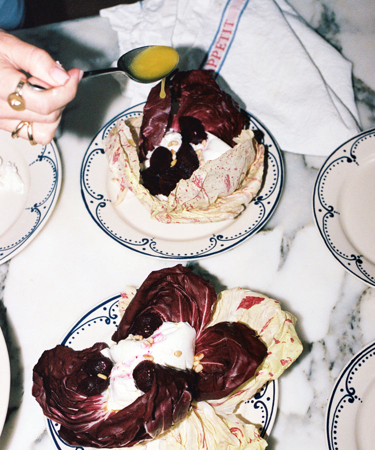 A hand drizzles yellow dressing over radicchio leaves filled with beets and cream, served on the Arco Small Plate, Set of 4, featuring a traditional design, placed on a marble surface with a napkin and other plates.