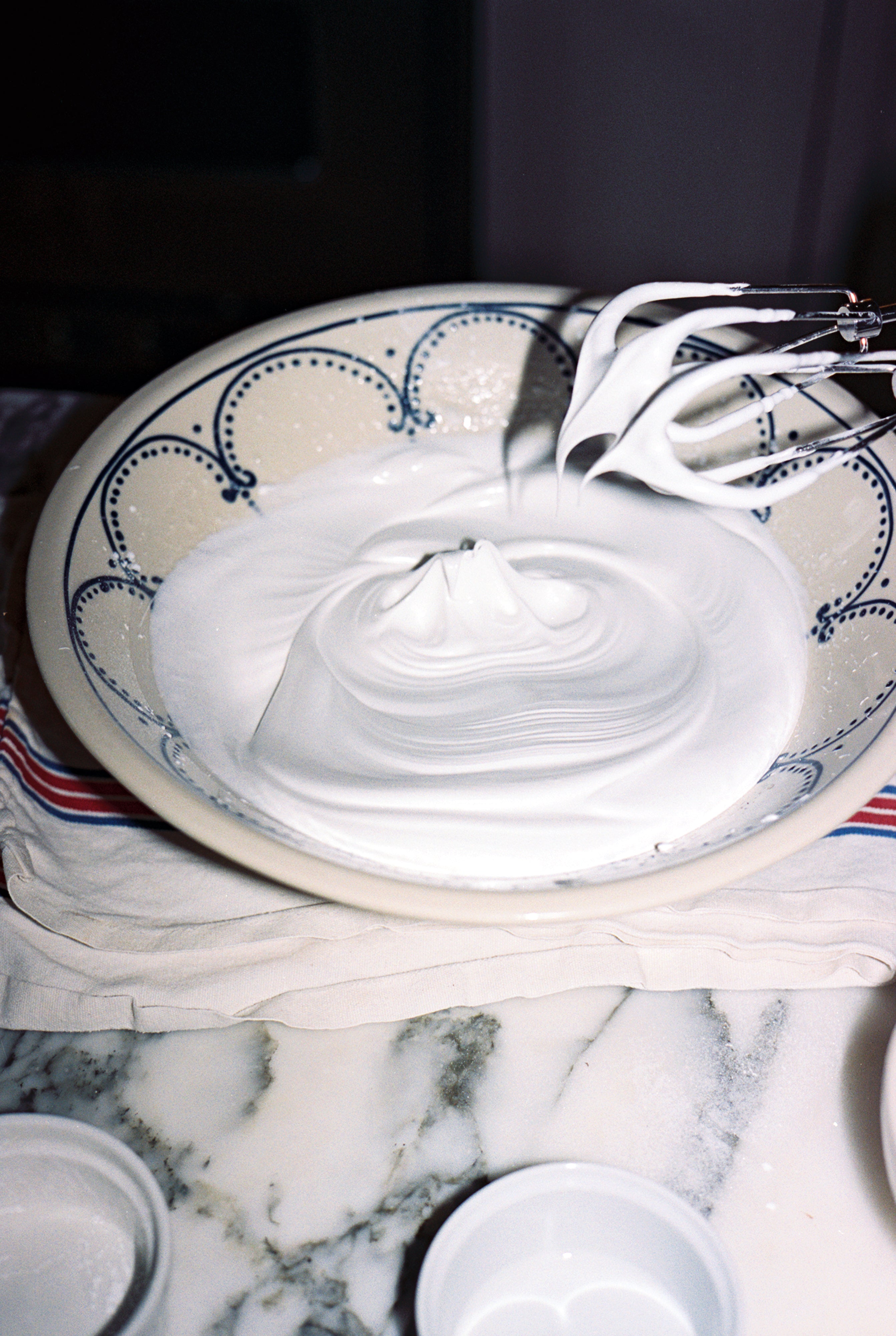 The handmade Arco Serving Bowl holds thick, glossy whipped egg whites on a kitchen counter, as a beater lifts out stiff peaks.