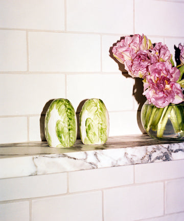 Two halves of a cabbage, a glass vase with pink flowers, and the Radicchio Salt and Pepper Shakers are displayed on a marble shelf against a tiled wall.