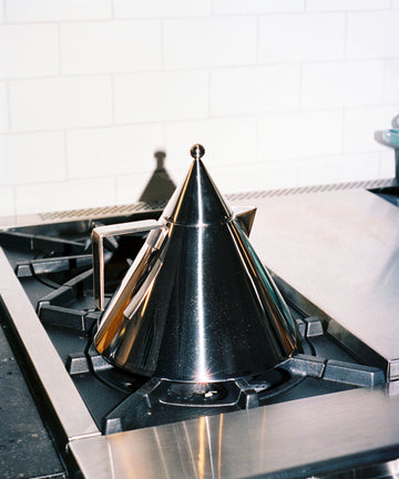 The Il Conico Kettle, crafted from stainless steel in a cone shape and inspired by Aldo Rossi, sits on a gas stove against a white subway tile backsplash, subtly referencing modernist design.