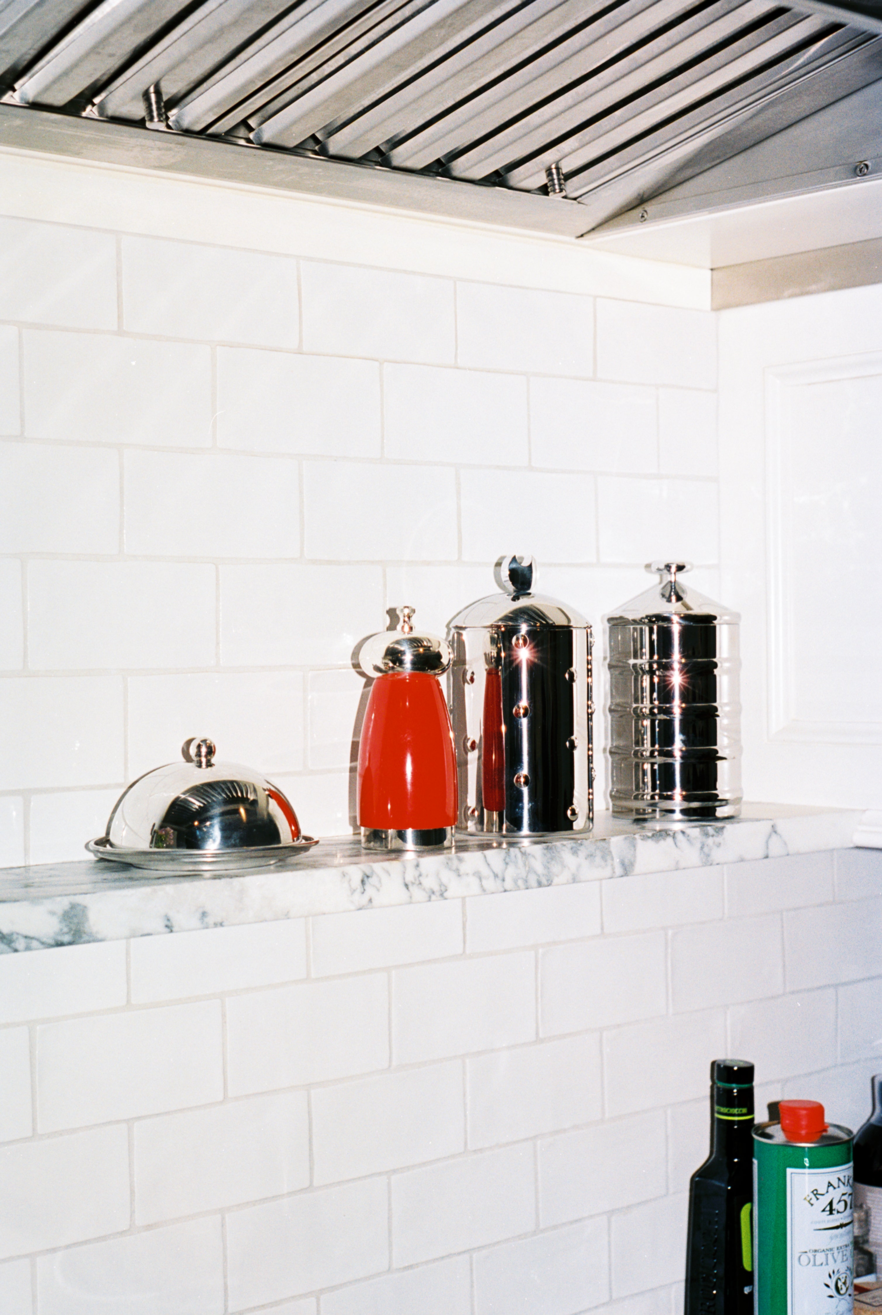 The Kalisto Kitchen Box, Italian-crafted metal containers including a red coffee pot and covered butter dish, sits on a marble shelf against white tile, with bottles of oil below.