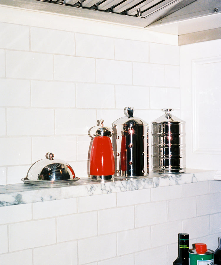The Kalisto Kitchen Box, Italian-crafted metal containers including a red coffee pot and covered butter dish, sits on a marble shelf against white tile, with bottles of oil below.