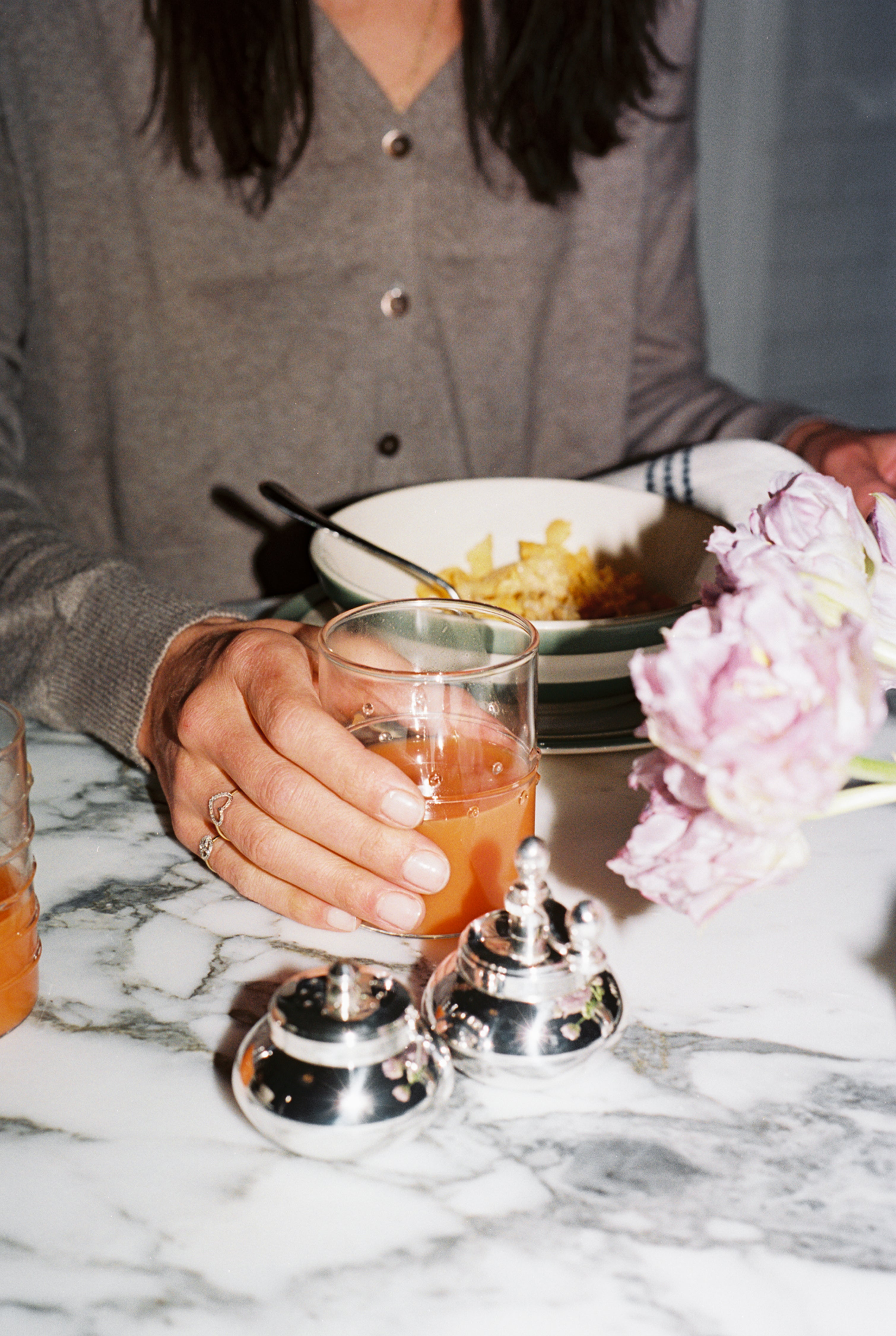 A person in a brown top sits at a marble table holding a glass from the Deco Water Glass, Set of 6, surrounded by vibrant flowers, a bowl of food, and silver condiment containers for an inviting tablescape.