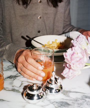 A person in a brown top sits at a marble table holding a glass from the Deco Water Glass, Set of 6, surrounded by vibrant flowers, a bowl of food, and silver condiment containers for an inviting tablescape.