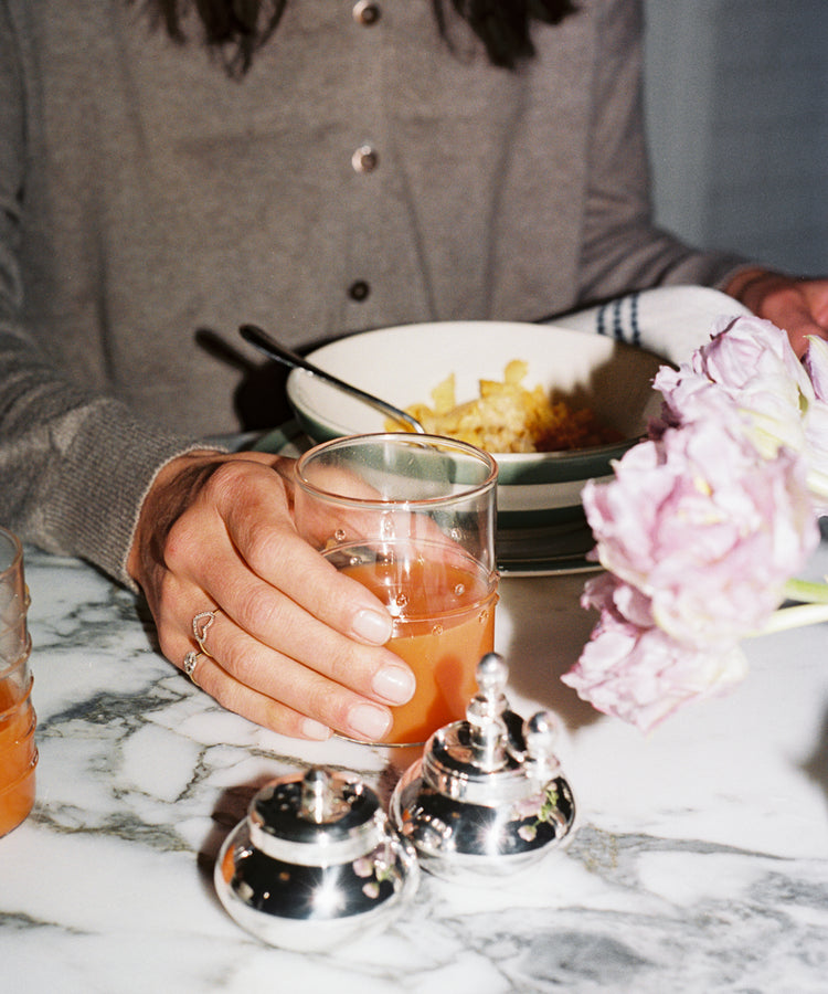 A person in a brown top sits at a marble table holding a glass from the Deco Water Glass, Set of 6, surrounded by vibrant flowers, a bowl of food, and silver condiment containers for an inviting tablescape.