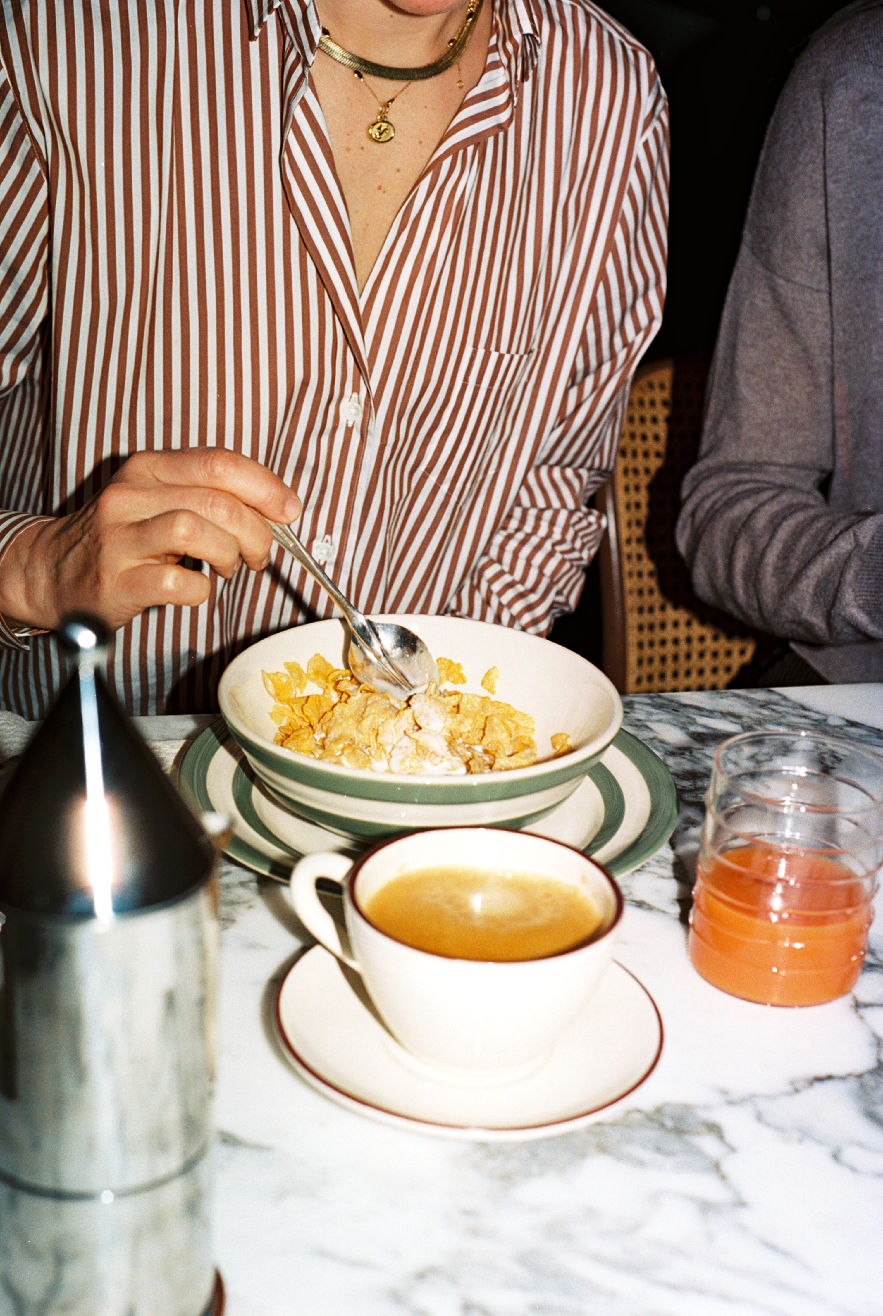 A person in a striped shirt enjoys breakfast at a marble table with the Betty Cereal Bowl, coffee, juice, and a metal container nearby. West Country pottery adds a charming touch to the setting.