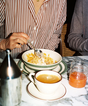 A person in a striped shirt enjoys breakfast at a marble table with the Betty Cereal Bowl, coffee, juice, and a metal container nearby. West Country pottery adds a charming touch to the setting.
