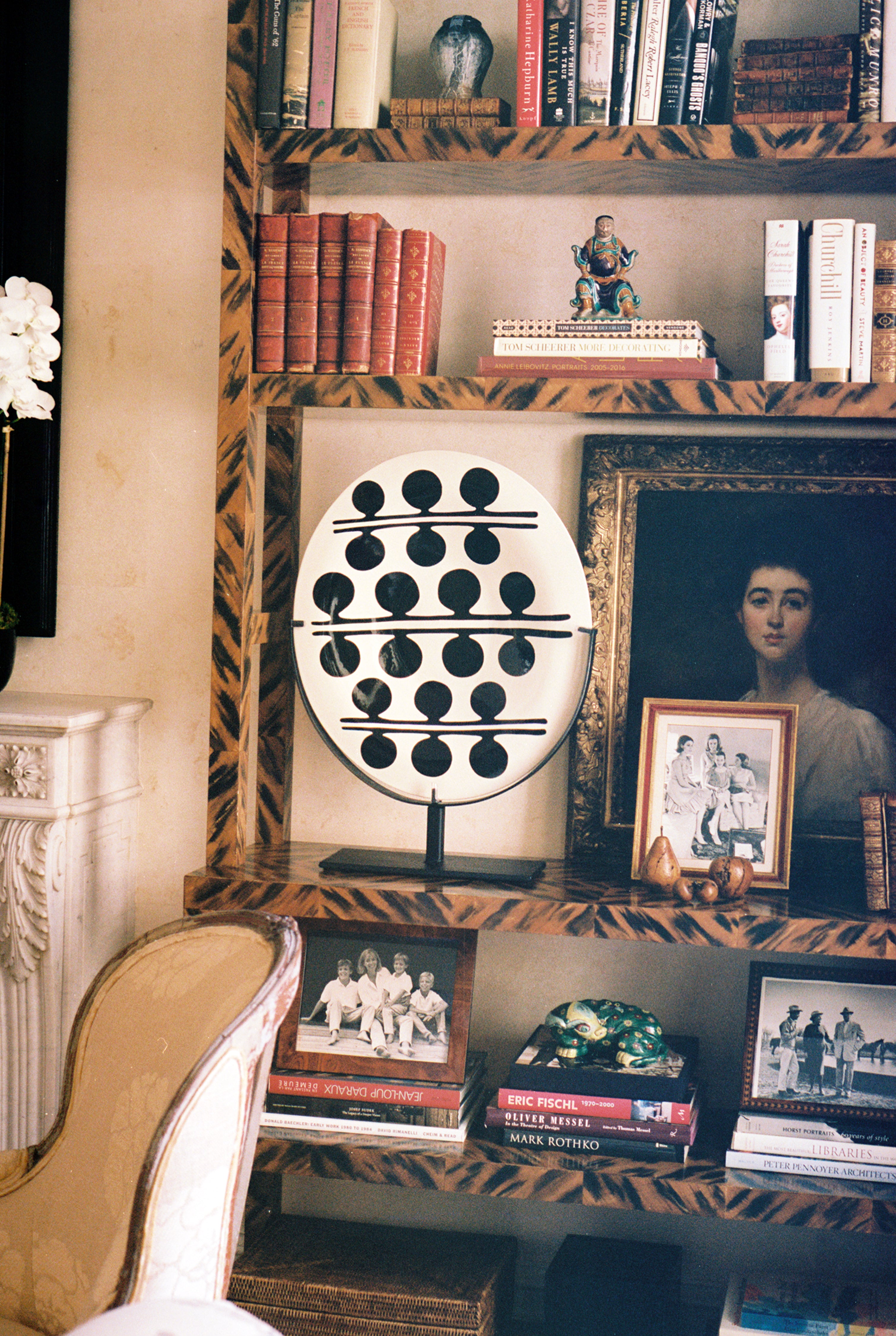 The Motif 3, Oval is a handcrafted glazed earthenware platter displayed on a wooden bookshelf alongside books, framed family photos, a large portrait painting, a decorative orb sculpture, and other small décor items.