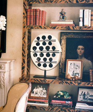 The Motif 3, Oval is a handcrafted glazed earthenware platter displayed on a wooden bookshelf alongside books, framed family photos, a large portrait painting, a decorative orb sculpture, and other small décor items.