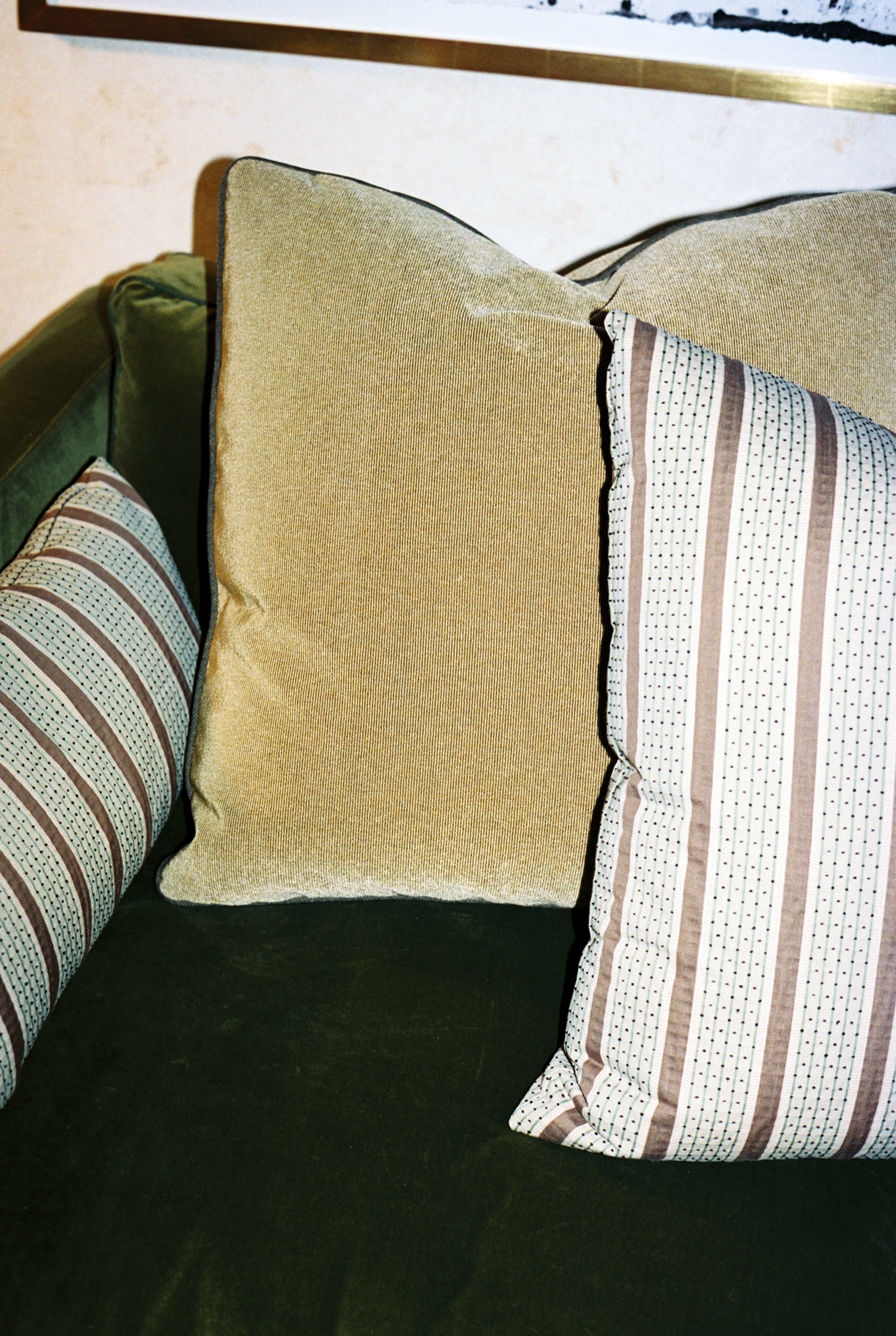 Two Marco Square Cushions made from premium deadstock designer fabrics—one in solid beige and one with stripes—are displayed on a green upholstered couch.