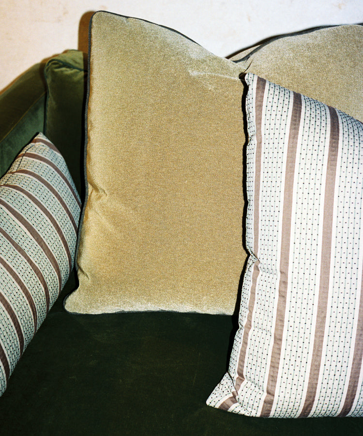 Two Marco Square Cushions made from premium deadstock designer fabrics—one in solid beige and one with stripes—are displayed on a green upholstered couch.