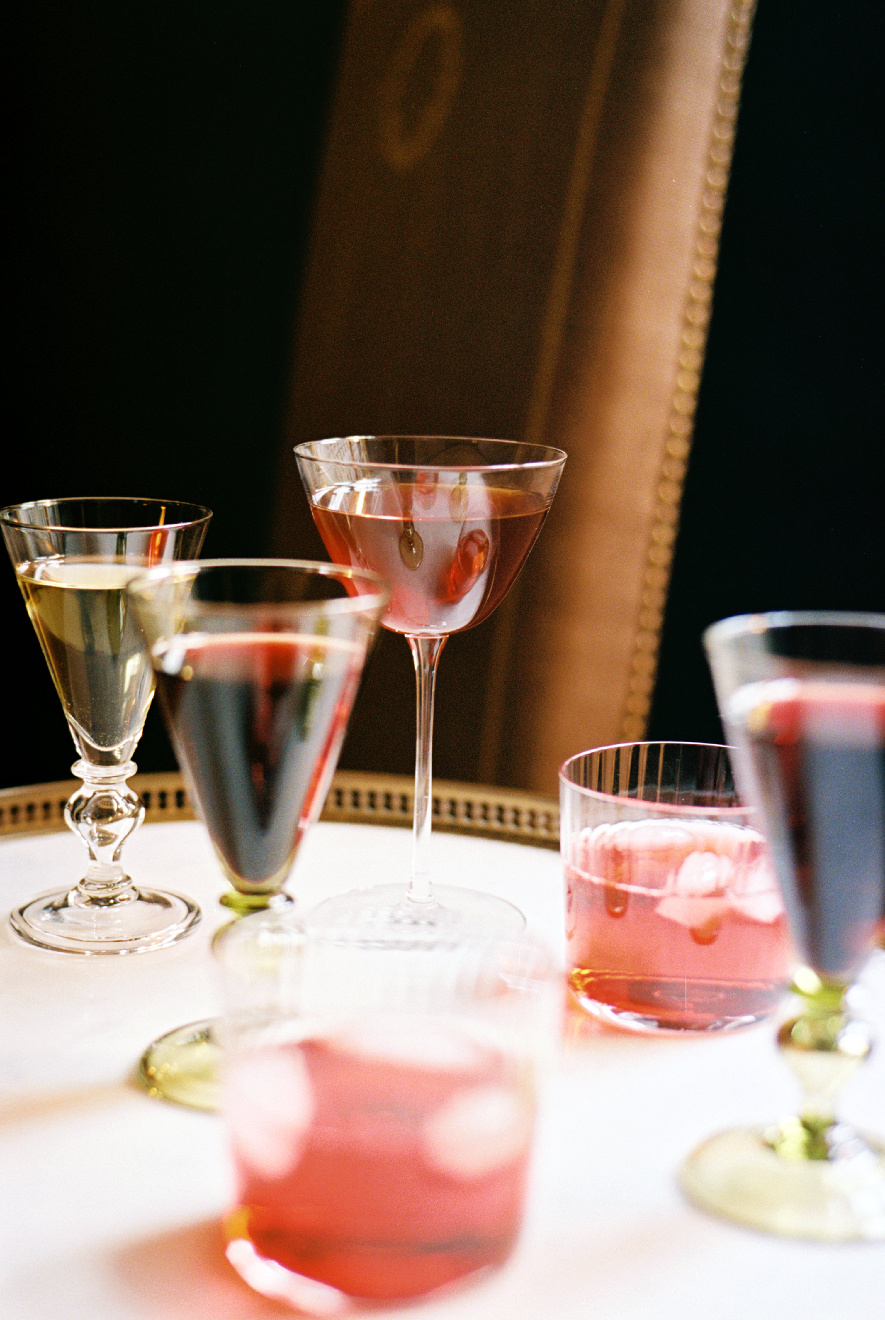 A variety of cocktails, including the Perfect Martini served in a Tina Martini Glass (Set of 4), are displayed on a round white table in front of a brown upholstered chair.