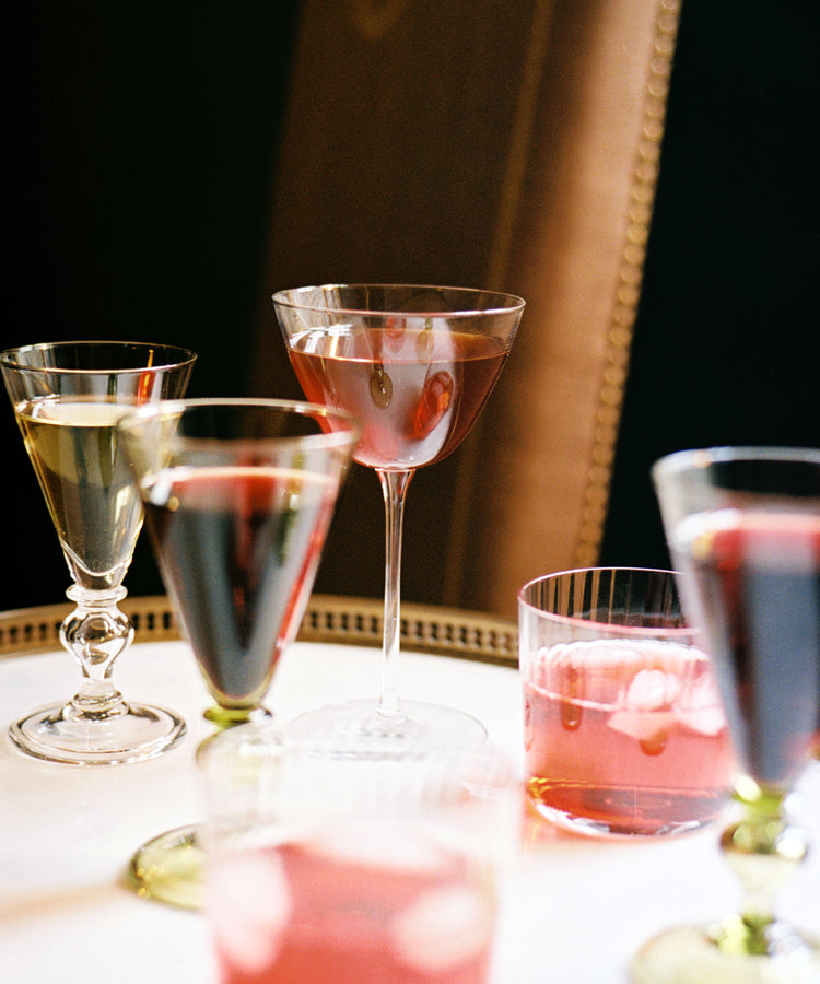 A variety of cocktails, including the Perfect Martini served in a Tina Martini Glass (Set of 4), are displayed on a round white table in front of a brown upholstered chair.