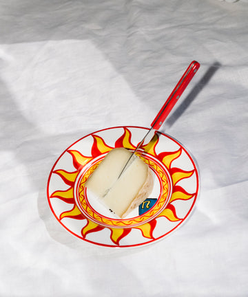 A large cheese knife with a red handle rests on a wedge of cheese atop a sun-patterned plate, all set against a white fabric background.